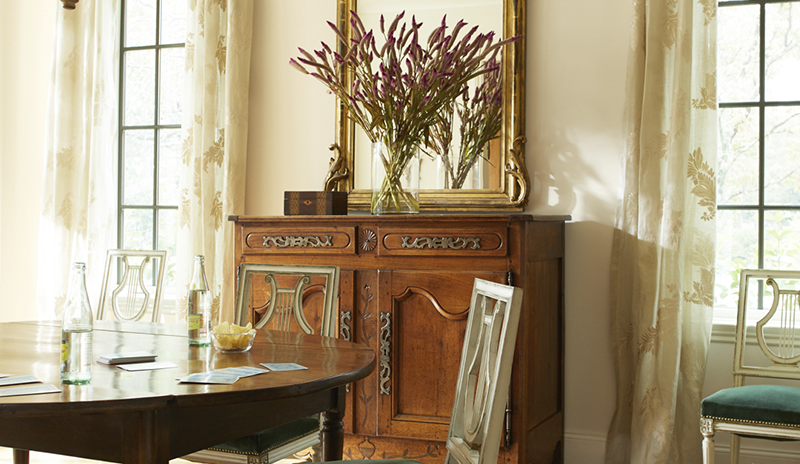 Elegant dining room with off-white walls, mixed wood furniture, gold accents, and beige drapes.