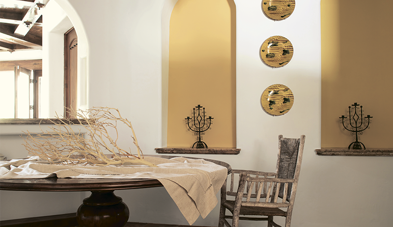 White dining room with yellow alcoves, plates, dark beams, rustic chandelier, and round wood table.